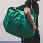 Mężczyzna niosący na ramieniu duży, zielony worek transportowy do tekstyliów używany w profesjonalnej logistyce pralniczej.